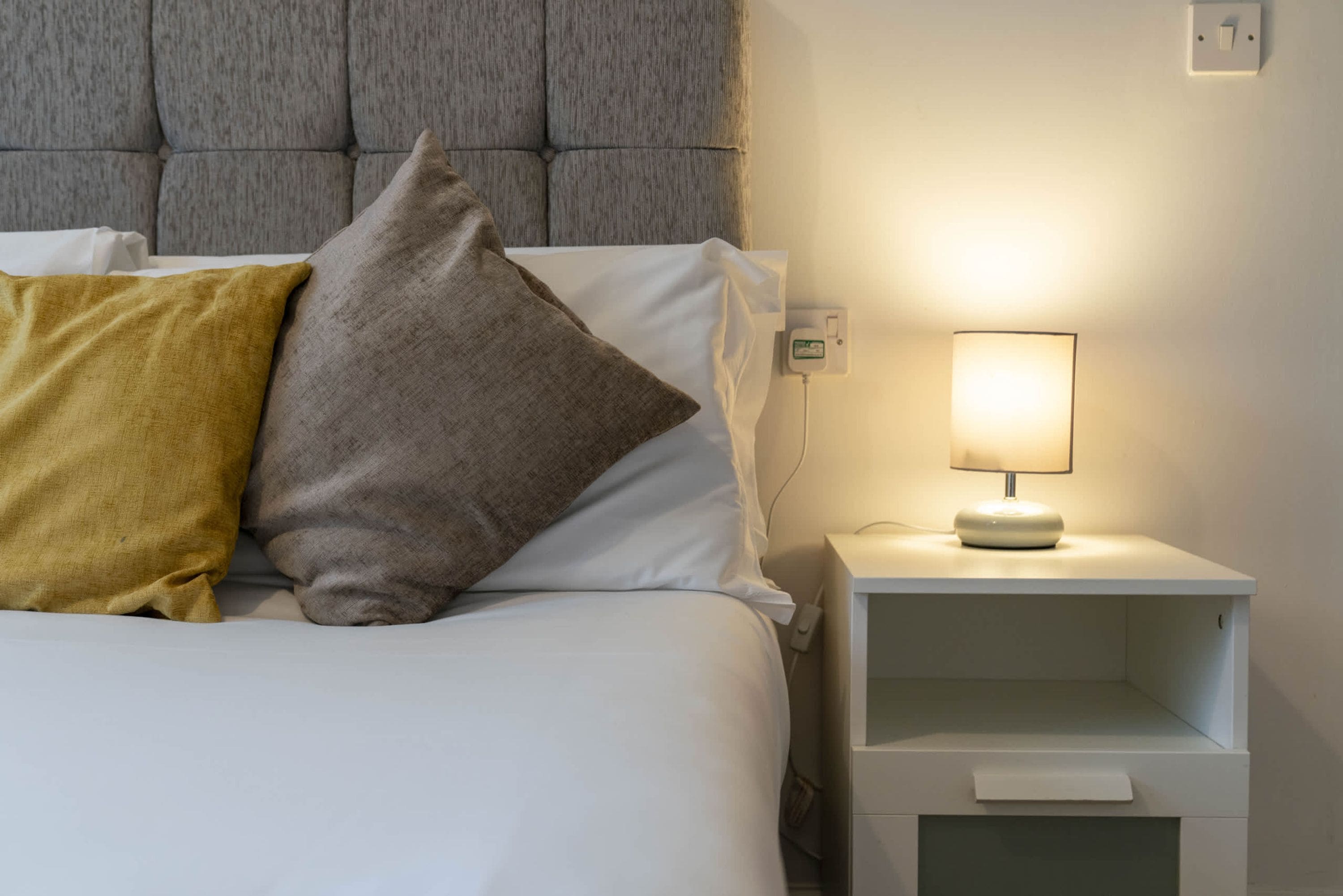 A neatly made bed with white bedding and two pillows, one yellow and one gray, against a gray headboard. A bedside table with a lamp and a power outlet is visible.