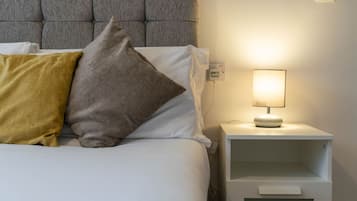A neatly made bed with white bedding and two pillows, one yellow and one gray, against a gray headboard. A bedside table with a lamp and a power outlet is visible.