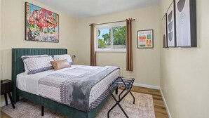 A bedroom with a bed, a window, curtains, a bedside table, a lamp, and wall art.