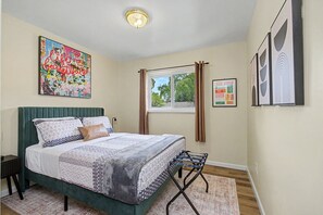A bedroom with a bed, a window, curtains, a bedside table, a lamp, and wall art.