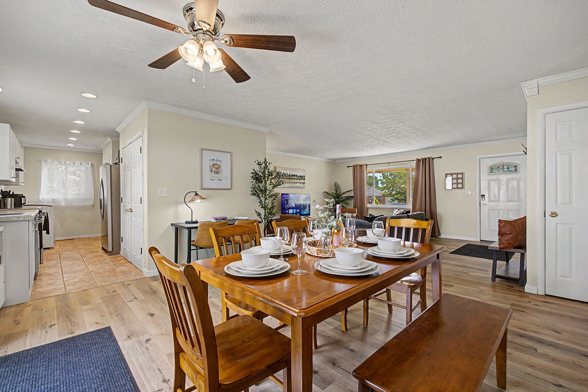 A dining area with a wooden table set for four, a ceiling fan, and a kitchen in the background.