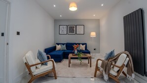 A modern living room with a blue sofa, two wooden chairs, a coffee table with a vase, and a black radiator. - Spacious 5bed House in Ruislip Sleeps 10 (Ruislip)