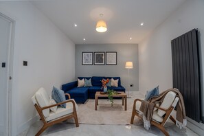 A modern living room with a blue sofa, two wooden chairs, a coffee table with a vase, and a black radiator. - Spacious 5bed House in Ruislip Sleeps 10 (Ruislip)