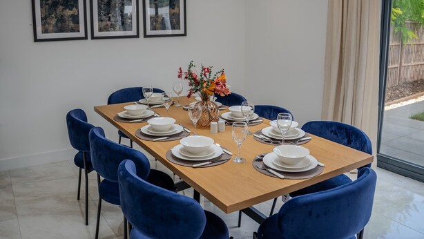 A dining area with a wooden table set for six, blue chairs, white plates, and clear glasses. There are three framed pictures on the wall above the table. - Spacious 5bed House in Ruislip Sleeps 10 (Ruislip)