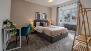 A bedroom with a bed, bedside tables, a desk, a chair, a window with a view, and a hanging light fixture. - Spacious 5bed House in Ruislip Sleeps 10 (Ruislip)