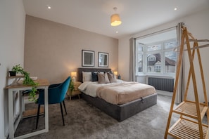 A bedroom with a bed, bedside tables, a desk, a chair, a window with a view, and a hanging light fixture. - Spacious 5bed House in Ruislip Sleeps 10 (Ruislip)
