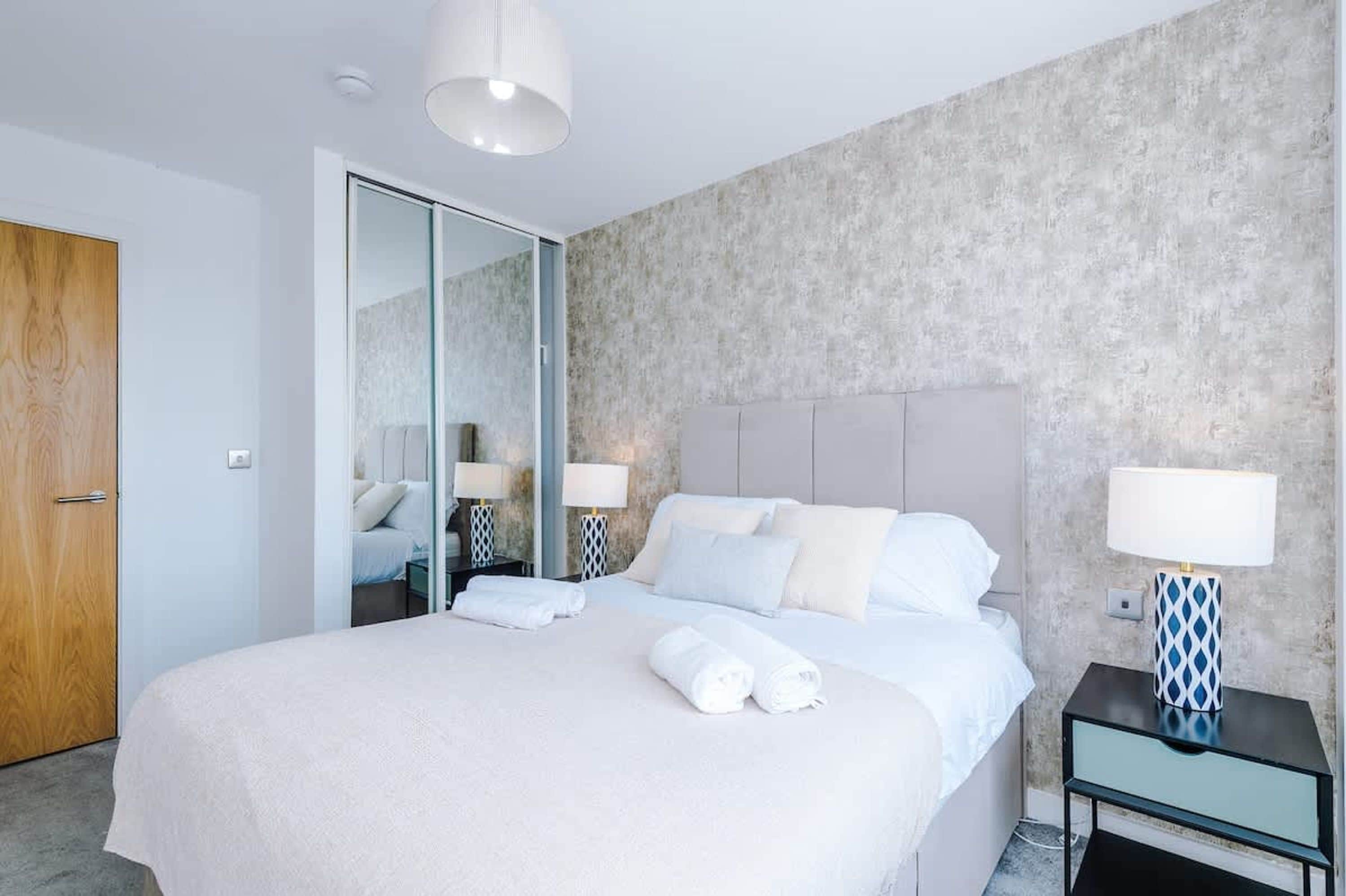 A modern hotel room with a large bed, a mirror, a bedside table with a lamp, and a wooden door.