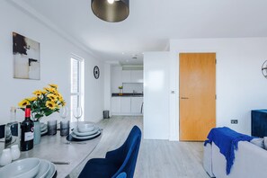A modern kitchen with white cabinets, a wooden door, and a dining area with a blue chair and a table set for two. - Modern 2 Bed Duplex Free Parking Wi Fi (Salford)