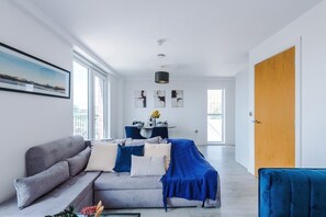 A modern living room with a sofa, a dining table, and a kitchen area in the background. - Modern 2 Bed Duplex Free Parking Wi Fi (Salford)
