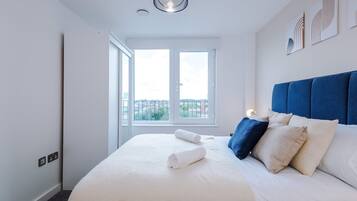 A hotel room with a large bed, a view through a window, and a blue headboard.