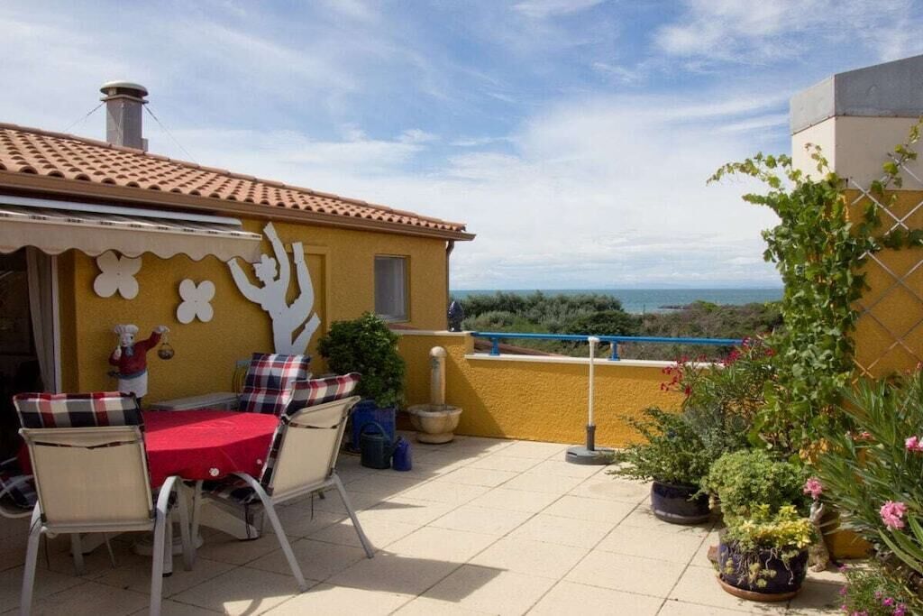 A patio with a table and chairs, a yellow wall with decorative elements, and a view of the sea.