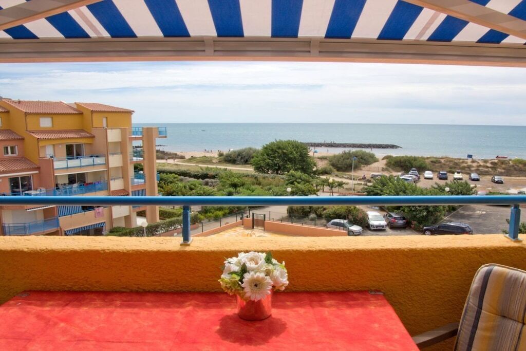 Un balcón con una mesa roja, un jarrón de flores y vistas al mar y edificios.