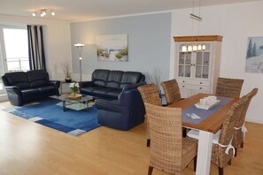 A living room with a blue sofa, a coffee table, and a dining area with wicker chairs and a wooden table. - Strandpalais, Whg. 127 Penthouse (Cuxhaven)