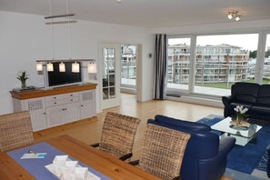 A modern living room with a dining area, a flat-screen TV, and a view of residential buildings. - Strandpalais, Whg. 127 Penthouse (Cuxhaven)