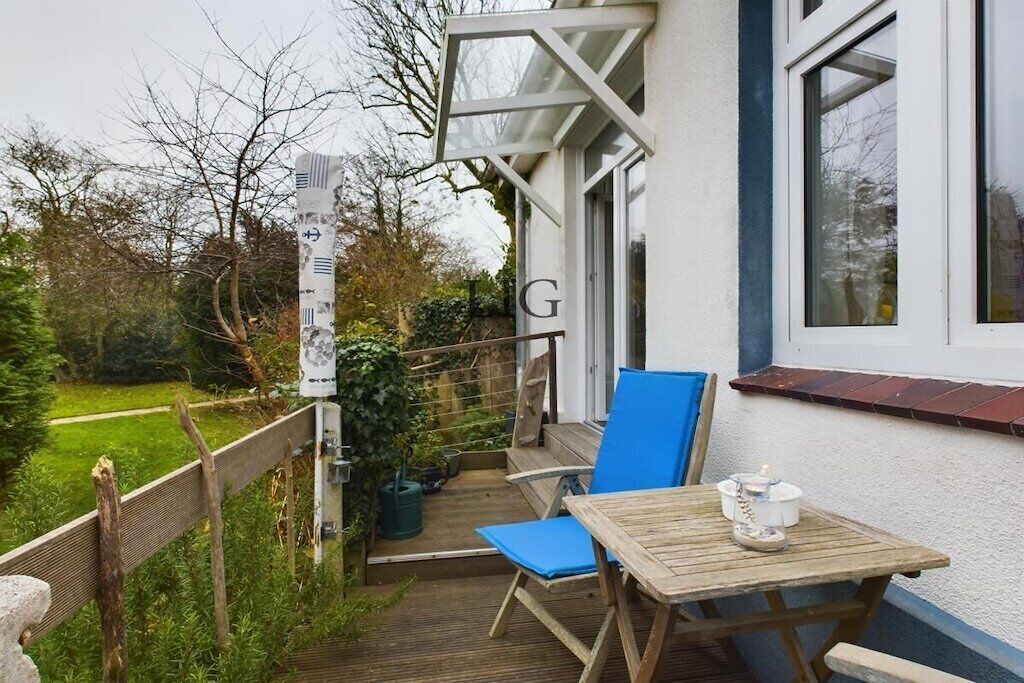 Eine Holzterrasse mit Tisch und Stühlen, ein kleiner Gartenbereich und ein weißes Gebäude mit einem gut sichtbaren Buchstaben 'G' an der Wand.