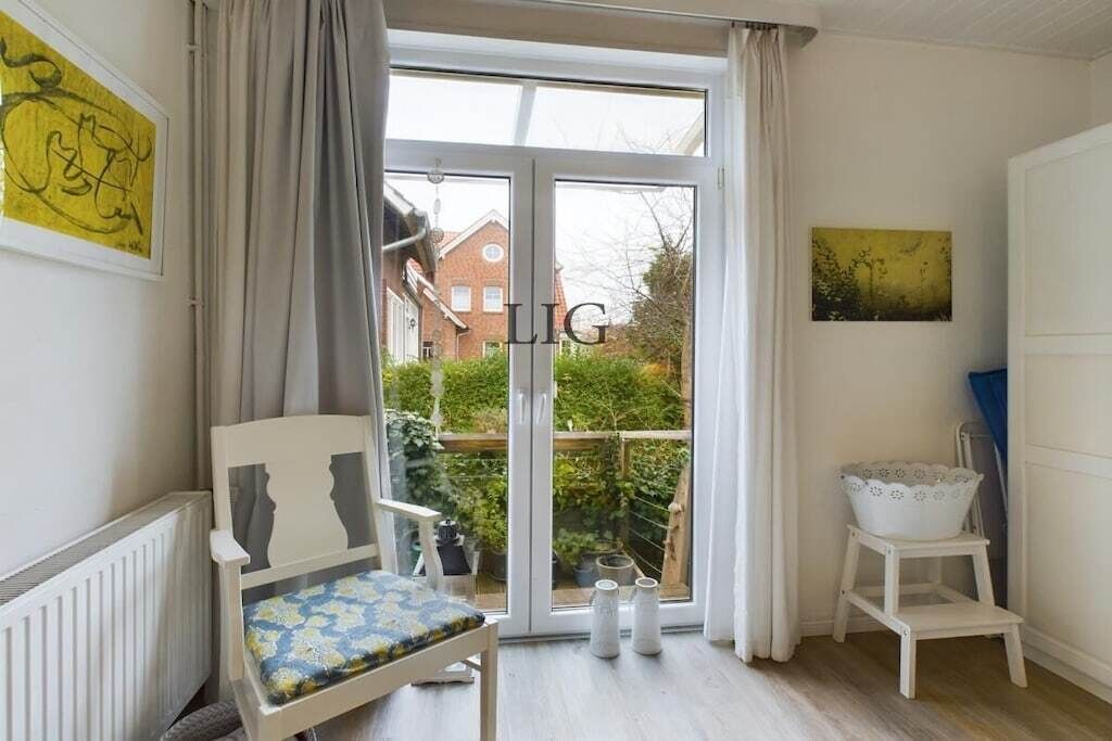 Ein Zimmer mit Stuhl, einem kleinen Tisch, einer Heizung, einem Fenster mit Vorhängen und Blick auf einen Garten.