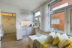 A compact hotel room with a kitchenette, a bed with pillows, a window with curtains, and a view of a brick building. - Holiday apartment cockle (Langeoog)