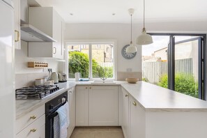 A modern kitchen with white cabinetry, a gas stove, a built-in oven, a window with a view of the outside, and a clock on the wall. - GuestReady - Charming London Home with Attic Room (London)
