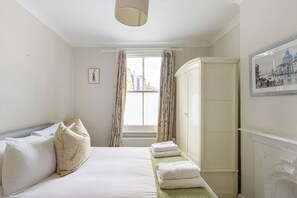 A hotel room with a bed, a wardrobe, a window with curtains, and a framed picture on the wall. - GuestReady - Charming London Home with Attic Room (London)