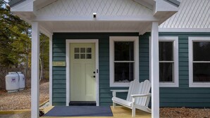 Une maison à l’extérieur vert, avec un porche blanc, une porte ornée d’une enseigne et deux chaises blanches.