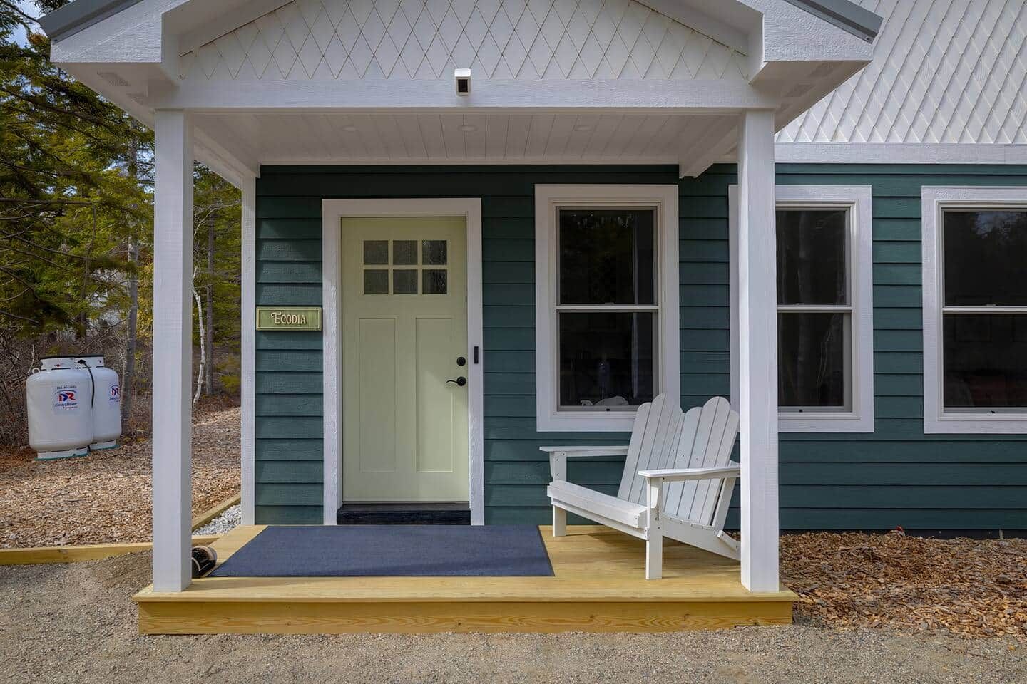 Une maison à l’extérieur vert, avec un porche blanc, une porte ornée d’une enseigne et deux chaises blanches.