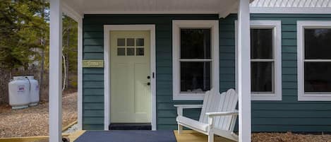 Una casa de color verde con un porche blanco, una puerta con un letrero y dos sillas blancas.