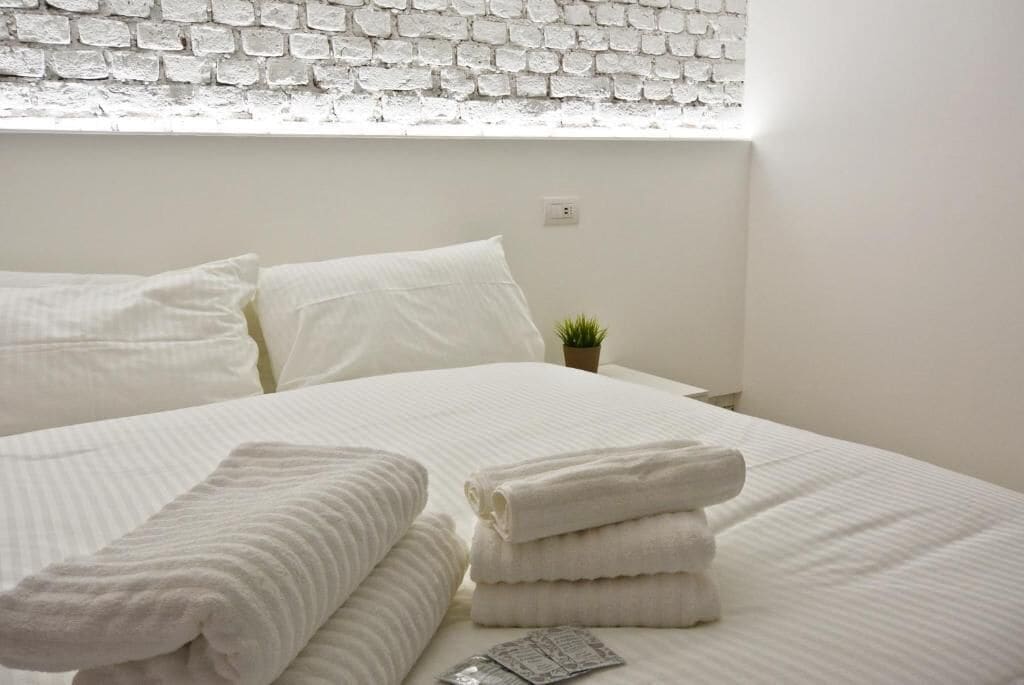 A neatly made bed with white linens and towels, a small potted plant on the nightstand, and a white brick wall.