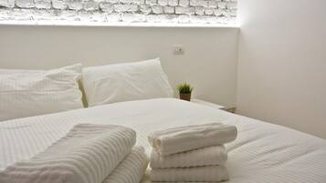 Una cama bien hecha con ropa de cama y toallas blancas, una pequeña planta en maceta sobre la mesita de noche y una pared de ladrillo blanco.