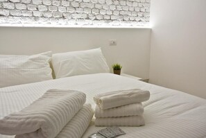 A neatly made bed with white linens and towels, a small potted plant on the nightstand, and a white brick wall. - Milano Apartments Lombardini (Milano)
