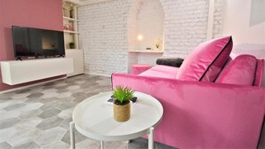 Ett vardagsrum med en vit tegelvägg, en rosa soffa, en platt-TV och ett litet runt bord med en växt.