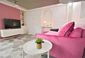 Ein Wohnzimmer mit weißer Ziegelwand, einem rosafarbenen Sofa, einem Flachbildfernseher und einem kleinen runden Tisch mit einer Pflanze.