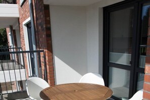 Een balkon met een ronde houten tafel en witte stoelen, een bakstenen muur en een glazen deur.