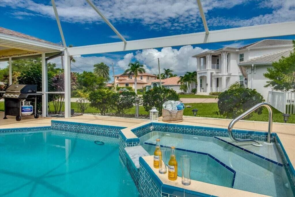 A swimming pool with a tiled edge, a covered area with a grill, and a view of a residential area with houses and greenery.