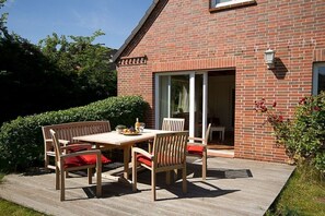Una casa in mattoni con un patio in legno, un tavolo e sedie, e una porta vetrata che dà su un ambiente interno.
