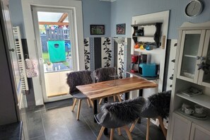 A dining area with a wooden table and chairs, a kitchen area with shelves and appliances, and a view of a backyard with a play structure. - Holiday apartment with 3 bedrooms (Langelsheim)