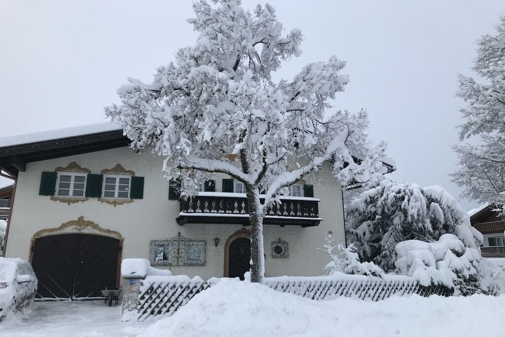 Um edifício coberto de neve com varanda, uma árvore na frente e uma cerca também coberta de neve.