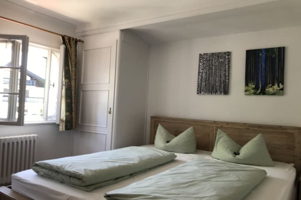 A bedroom with a bed, a radiator, a window with curtains, and two wall-mounted artworks.