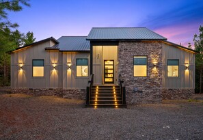 A modern two-story house with a stone facade and a metal roof, featuring a welcoming entrance with steps and illuminated exterior lights. - EV Charger | Hot Tub | Games | Sleeps 16 (Broken Bow)