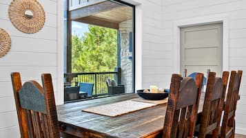 A wooden dining table with chairs, a woven wall decoration, and a view of greenery outside.