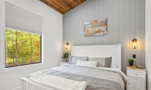 A bedroom with a large bed, two bedside tables, a window with a view of trees, and a wall-mounted light fixture. - EV Charger | Hot Tub | Games | Sleeps 16 (Broken Bow)