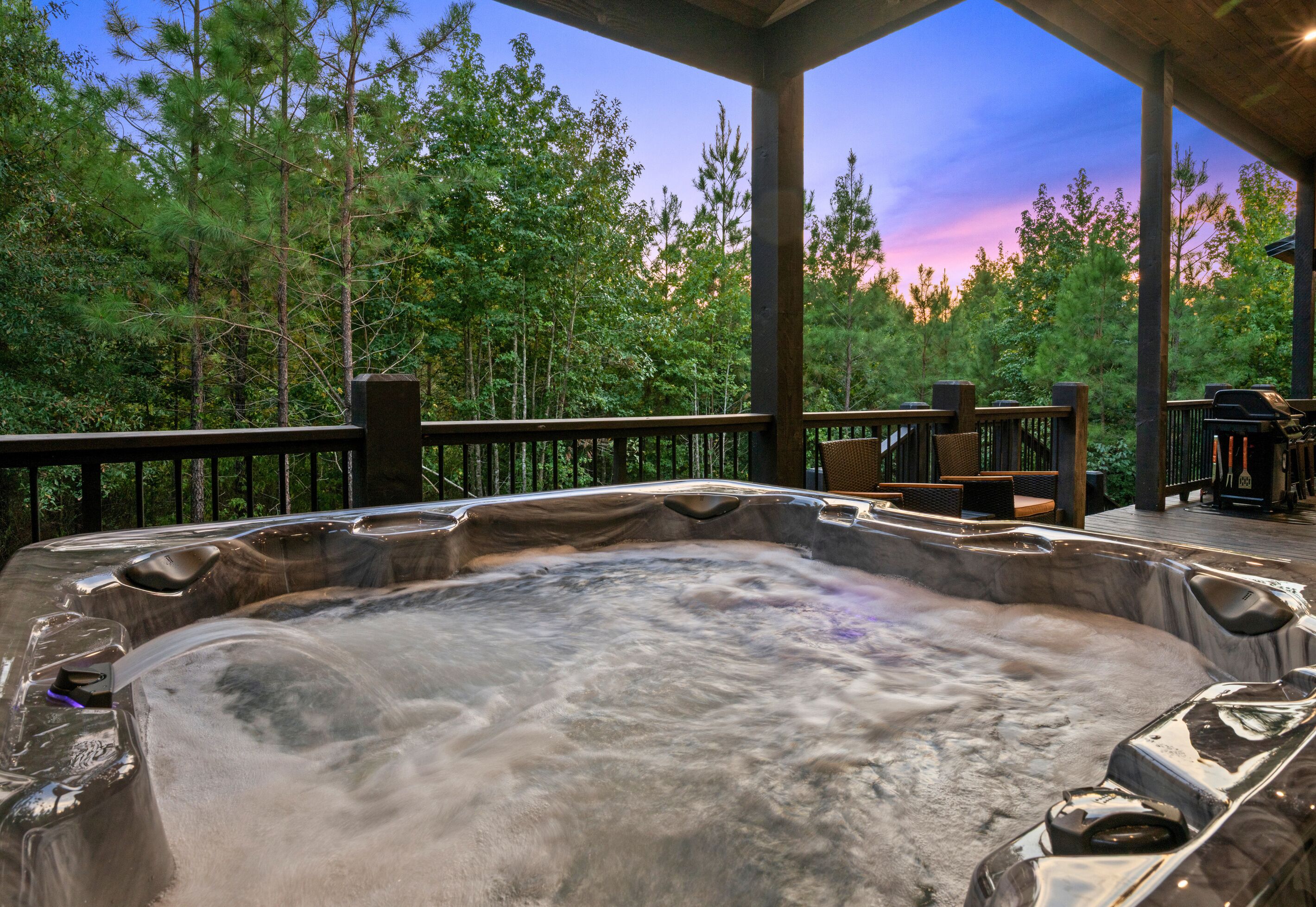 A hot tub on a wooden deck with a view of a forest and a sunset.