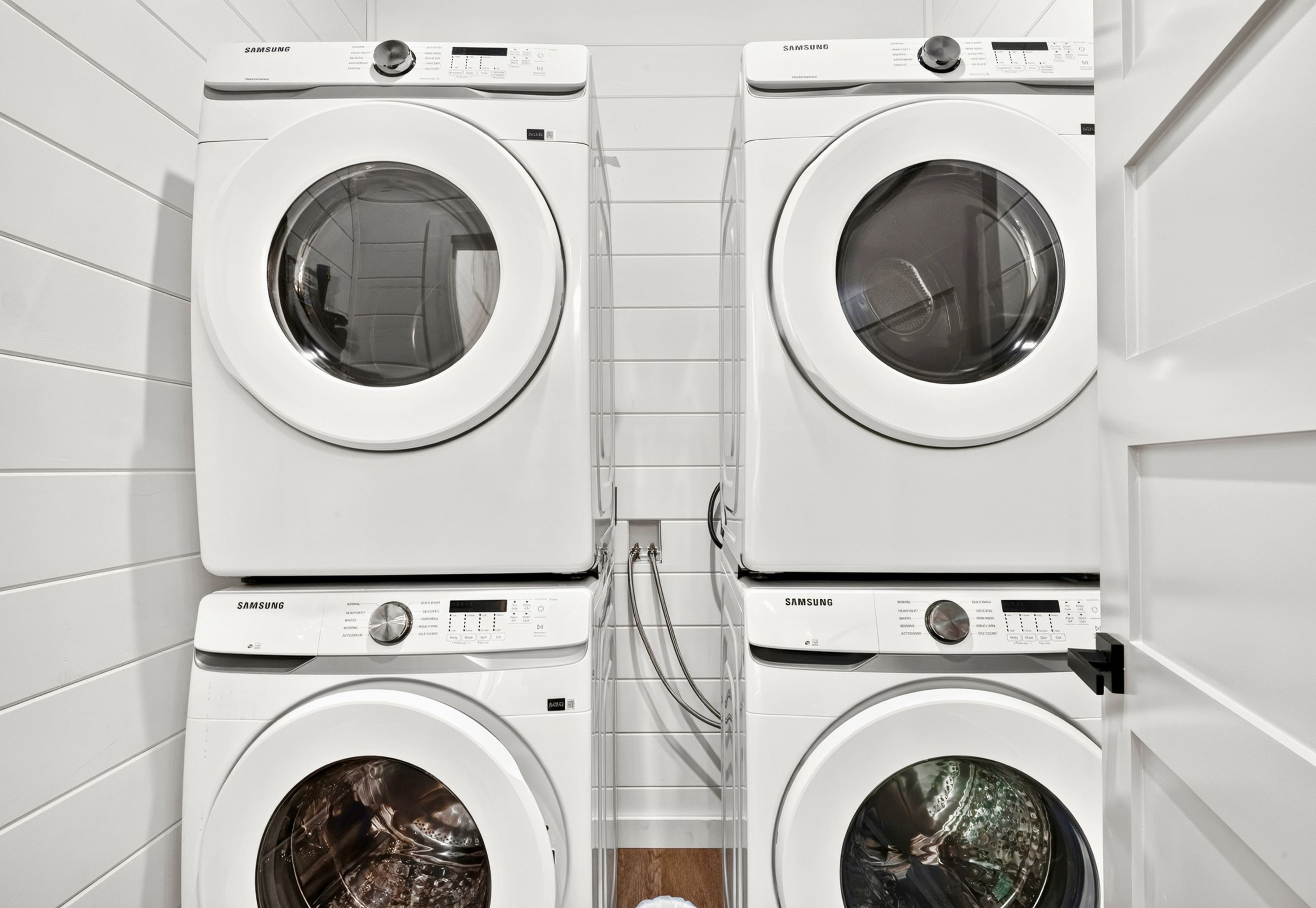 A laundry room with four white Samsung washing machines stacked in pairs against a tiled wall.