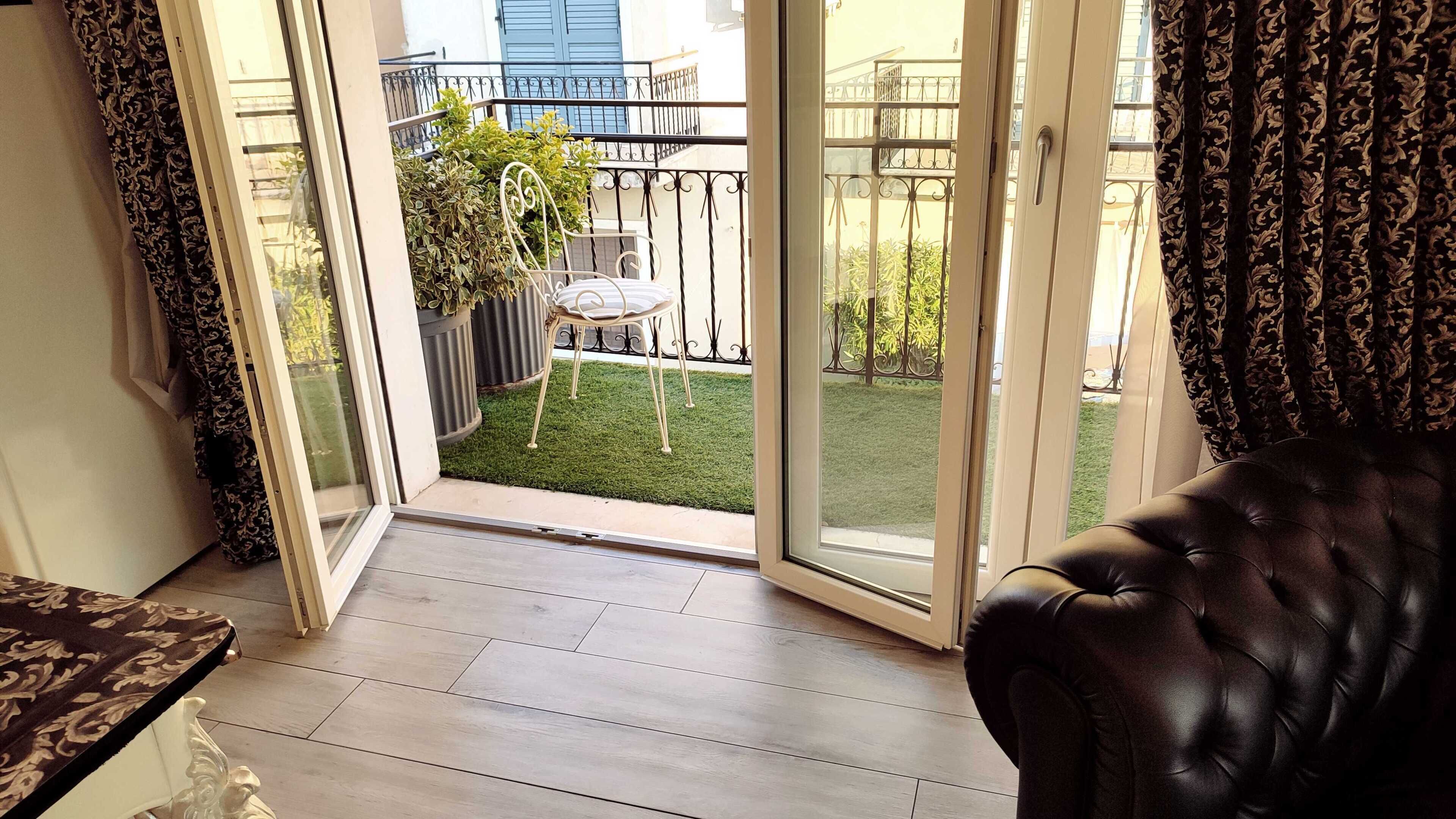 Una habitación con puerta corrediza de vidrio que da a un balcón con una pequeña mesa, una silla, una planta en maceta y un césped cuidado.