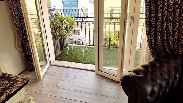 A room with a sliding glass door leading to a balcony with a small table and chair, a potted plant, and a neatly trimmed lawn.