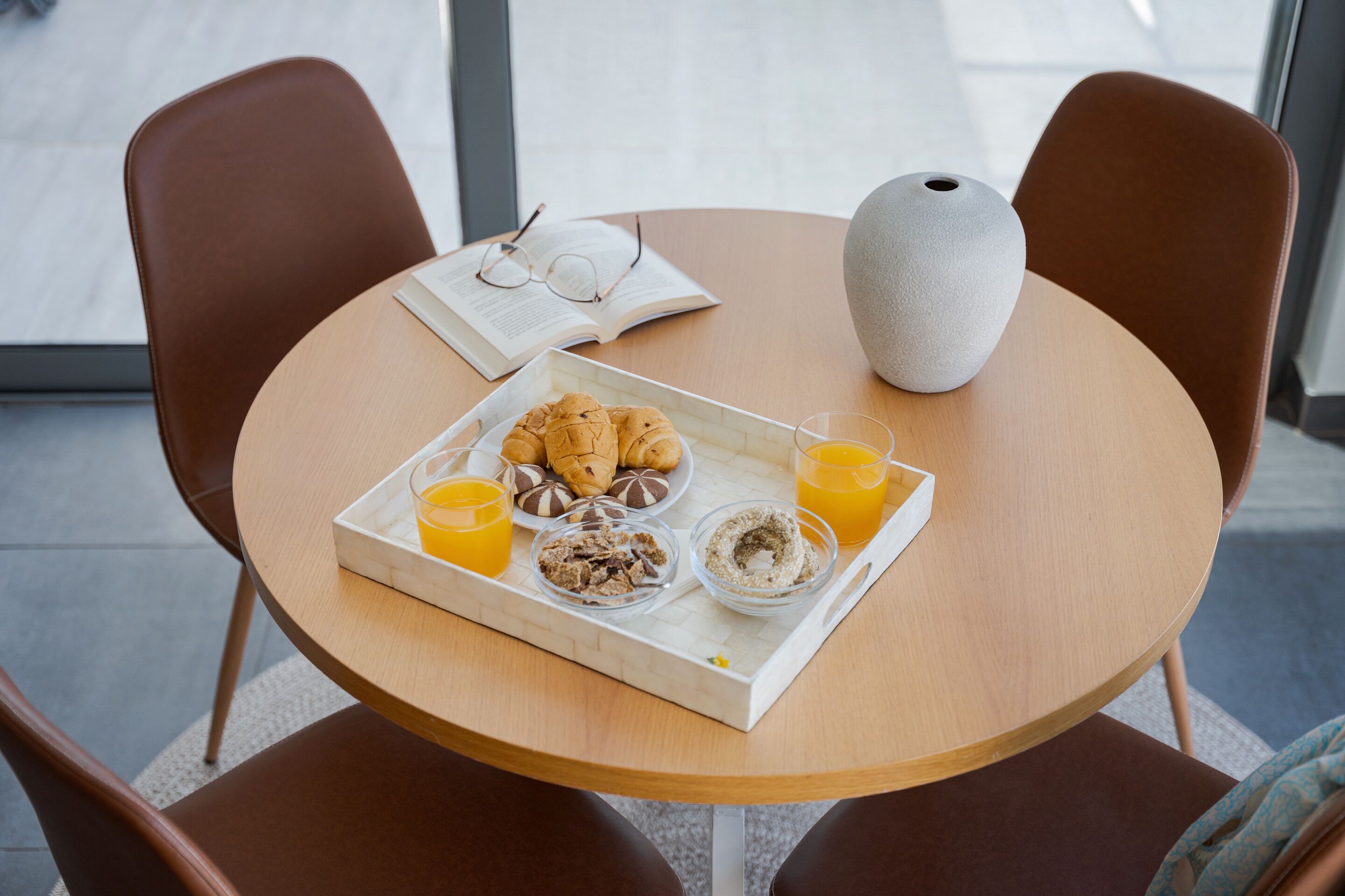 Ein runder Holztisch mit einer Tablett voller Frühstücksartikel, darunter Croissants, Müsli und Saft, sowie einer Vase auf dem Tisch.