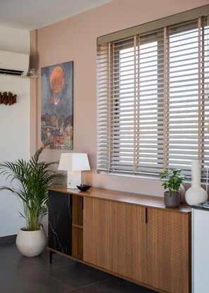 A modern interior with a wooden console, a potted plant, a wall-mounted lamp, and a window with blinds. - Aelia Riviera Apartment (Vari)