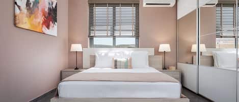 A modern bedroom with a large bed, two bedside tables, a window with blinds, an air conditioning unit, and a large mirror.