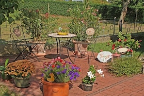 Uma área de pátio externa com mesa e cadeiras, rodeada por plantas e flores em vasos.