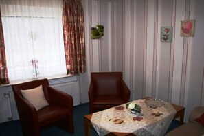 Ein Zimmer mit Fenster, zwei Stühlen, einem Tisch mit blumigem Tischläufer und einer Heizung.