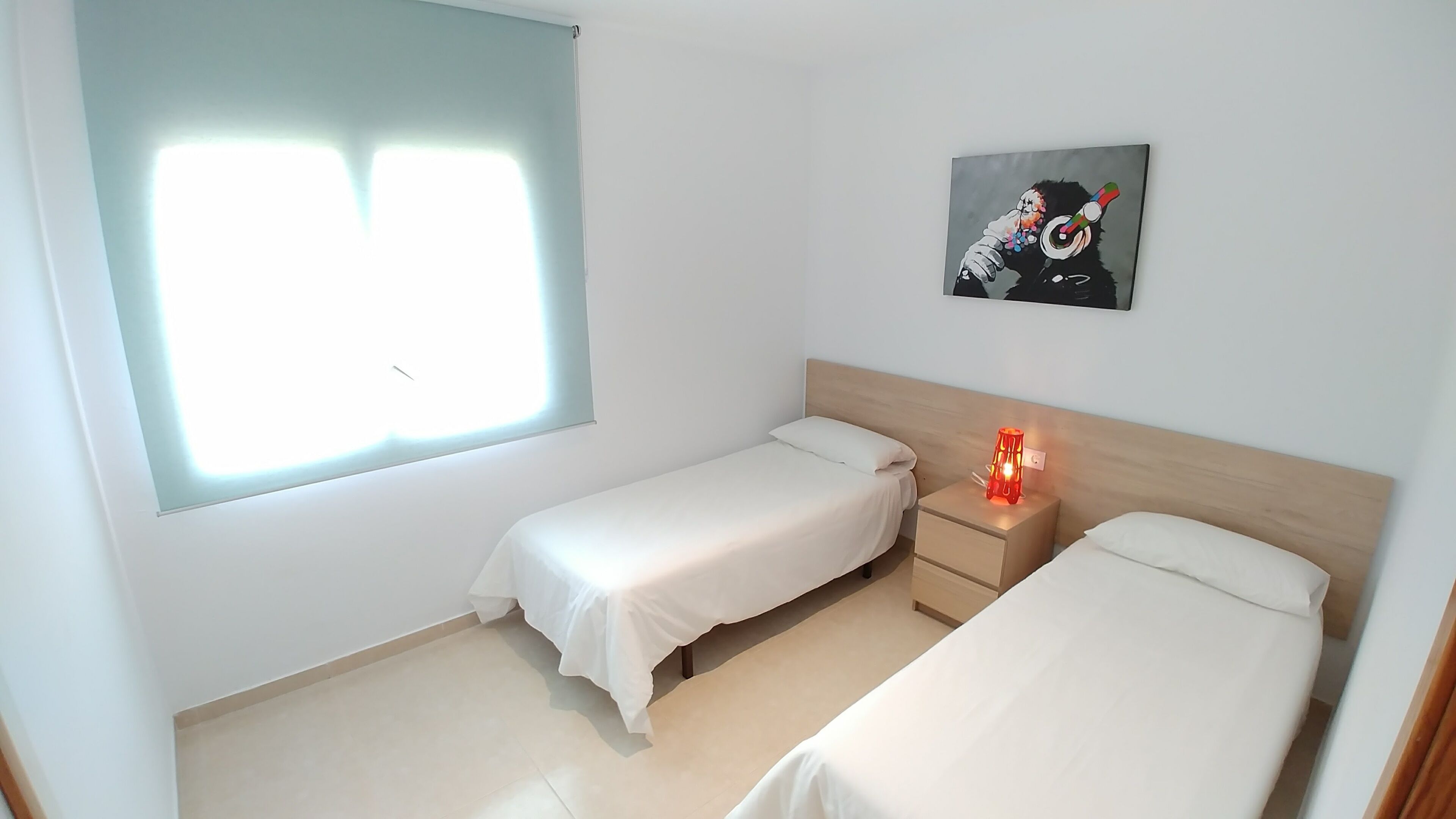 Una habitación con dos camas individuales, una mesita de noche con lámpara, una ventana con persianas y un cuadro en la pared.